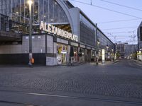 an empty, well - lit city street at dusk with people walking by the buildings