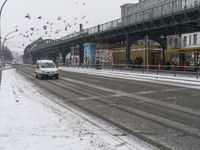 a city street covered in snow and lots of birds flying over it, next to a bridge with tracks