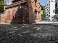 this is an old brick building next to a park with trees in the background and cobblestones in the foreground