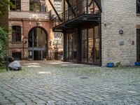 a bench and walkway at the side of a building with open windows, brick floors and doors