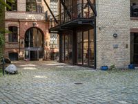 a bench and walkway at the side of a building with open windows, brick floors and doors