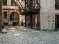 a bench and walkway at the side of a building with open windows, brick floors and doors