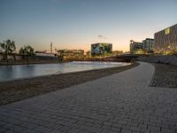 an empty brick pathway is next to a river and some buildings near it at night