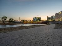 an empty brick pathway is next to a river and some buildings near it at night