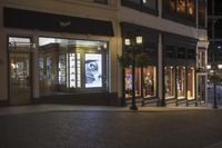 shopfronts are illuminated at night in an upscale mall setting with cobblestone walkway