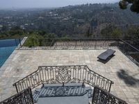 a balcony with a stone deck with a pool in the background, overlooking hills and a tree