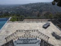 a balcony with a stone deck with a pool in the background, overlooking hills and a tree