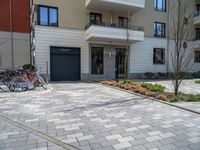 a row of bicycles parked on a brick road in front of an apartment building, with parking space in the background