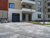 a row of bicycles parked on a brick road in front of an apartment building, with parking space in the background