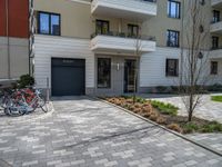 a row of bicycles parked on a brick road in front of an apartment building, with parking space in the background