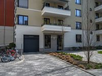 a row of bicycles parked on a brick road in front of an apartment building, with parking space in the background