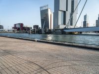 people ride bikes on a path over a river towards some city buildings with skyscrapers