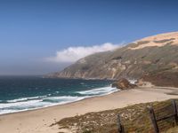 the sandy beach and ocean are along the coast line with rocky cliffs and clouds above