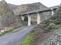 a bridge crosses an overpass above a road surrounded by mountains and trees, with low shrubs