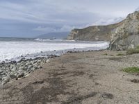 a beach near a cliff next to the ocean and rocks in the sand below a cloudy sky