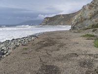 a beach near a cliff next to the ocean and rocks in the sand below a cloudy sky