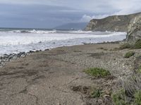 a beach near a cliff next to the ocean and rocks in the sand below a cloudy sky