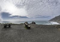 a fisheye lens captures three empty benches along the ocean edge with some big rocks behind