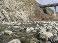 rocky river bed in mountains with water flowing under an overpass bridge and blue sky with clouds above