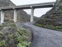 an overpass crossing over a mountain highway in the mountains below a bridge that overlooks a valley with shrubs and a small hill behind it