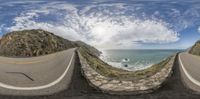 a wide angle view of a road near the ocean with some mountains behind it and sky in front of them