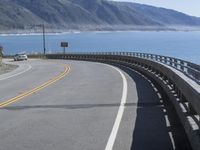 a curve along the ocean on a deserted road near mountains with waves coming in at the shore