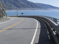 a curve along the ocean on a deserted road near mountains with waves coming in at the shore