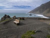 an empty bench on top of a mountain near the ocean near the beach, with large cliffs behind