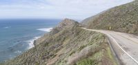 the road is winding along a side of a mountain by the ocean and blue sky