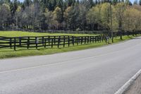 a black horse standing by the side of the road in front of a fenced in pasture