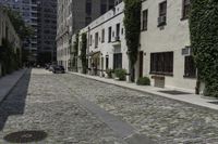 the cobblestone street has many stone sidewalks in front of it, where cars are parked and there is an outside bench with no visible