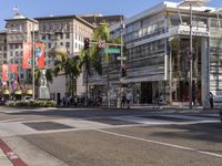 a blue car is stopped at an intersection with palm trees in the foreground, and shops and people walking in the street