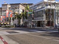 a blue car is stopped at an intersection with palm trees in the foreground, and shops and people walking in the street