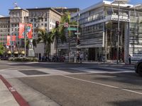 a blue car is stopped at an intersection with palm trees in the foreground, and shops and people walking in the street