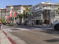 a blue car is stopped at an intersection with palm trees in the foreground, and shops and people walking in the street