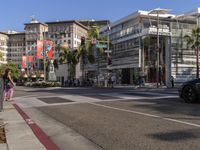 a blue car is stopped at an intersection with palm trees in the foreground, and shops and people walking in the street