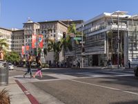 a blue car is stopped at an intersection with palm trees in the foreground, and shops and people walking in the street