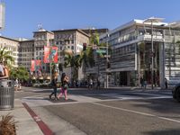 a blue car is stopped at an intersection with palm trees in the foreground, and shops and people walking in the street