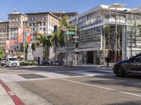 a blue car is stopped at an intersection with palm trees in the foreground, and shops and people walking in the street