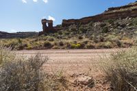 a dirt road leading to tall rocks and plants in the middle of the desert with trees and grass in front