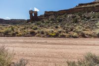 a dirt road leading to tall rocks and plants in the middle of the desert with trees and grass in front