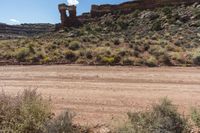 a dirt road leading to tall rocks and plants in the middle of the desert with trees and grass in front