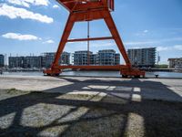 the red crane is on top of a dock by the water and buildings behind it