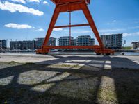 the red crane is on top of a dock by the water and buildings behind it