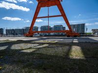 the red crane is on top of a dock by the water and buildings behind it