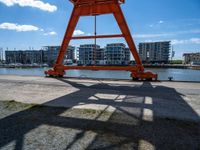 the red crane is on top of a dock by the water and buildings behind it