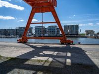 the red crane is on top of a dock by the water and buildings behind it