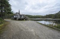 a boat with a ramp sitting in the gravel next to water in the woods while cloudy sky above