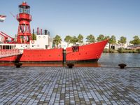 the large boat is sitting next to the light house and harbor tugboats on brick path near the river