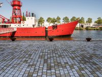 the large boat is sitting next to the light house and harbor tugboats on brick path near the river